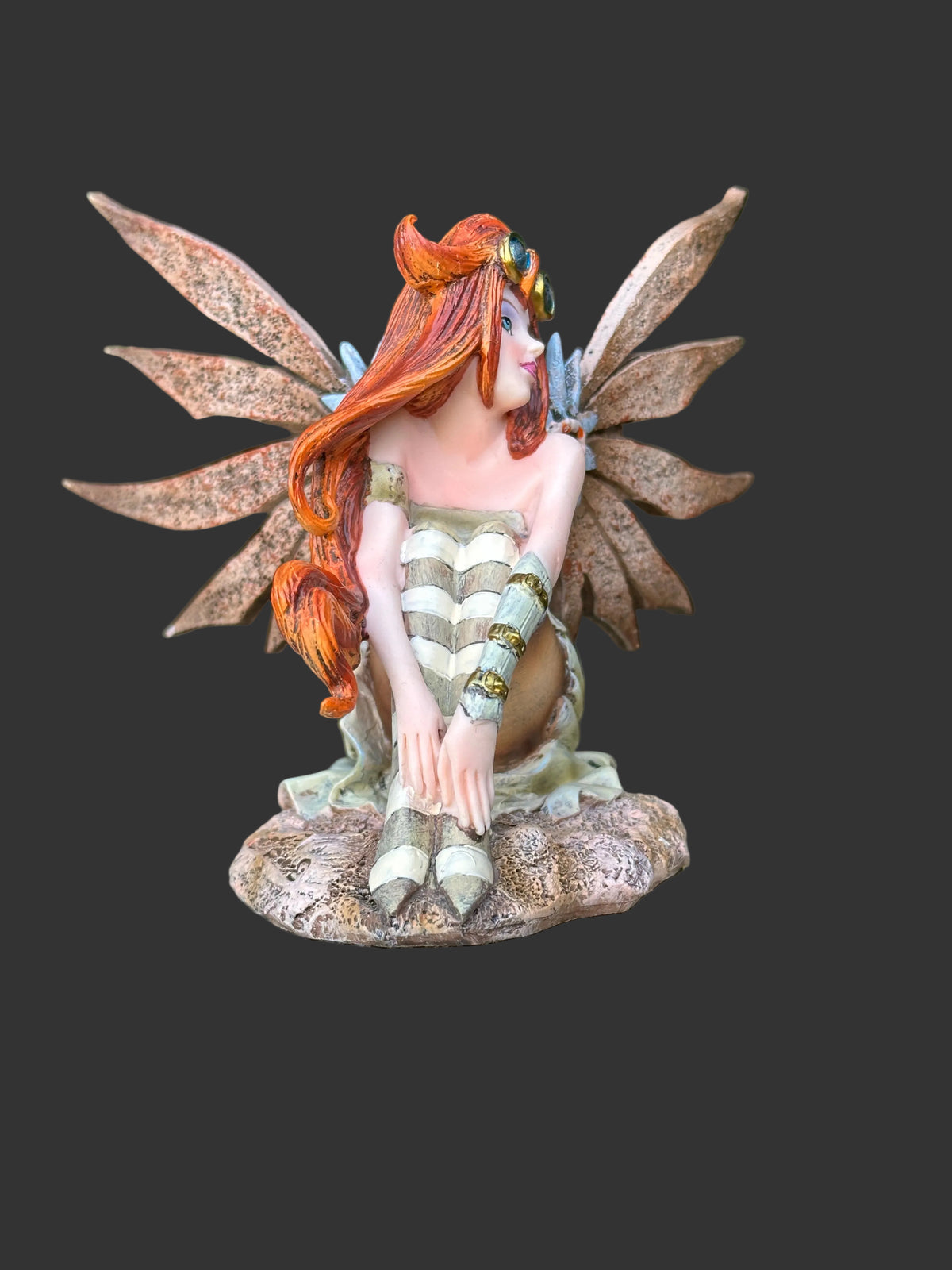 A detailed fantasy Amy Brown steampunk fairy figurine with orange hair, brown wings, and a green-and-white striped dress, sitting on a tree stump with a blurred garden background.