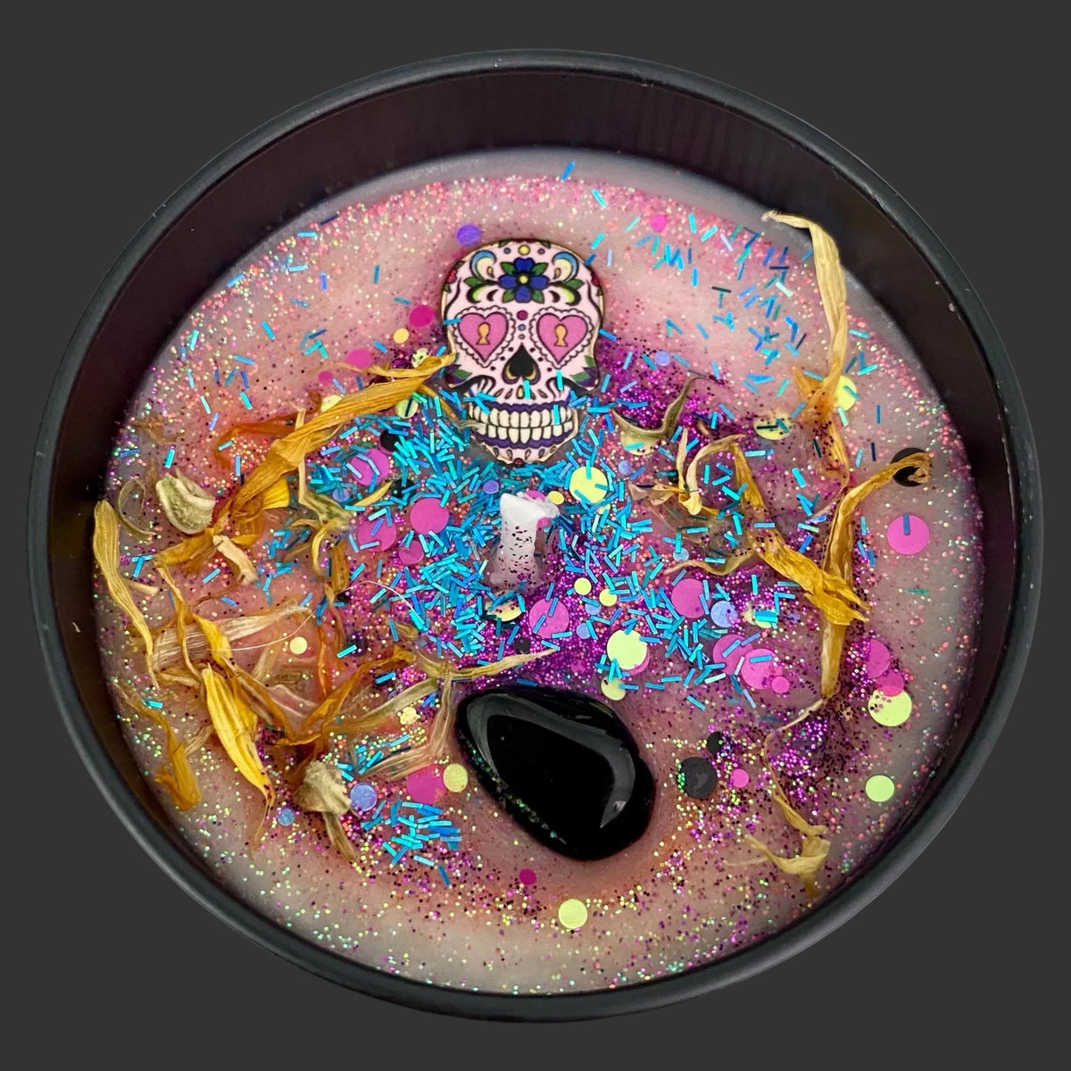 A round black candle with a pink wax base decorated for Dia de los Muertos, featuring a sugar skull ornament, dried yellow flowers, blue and pink glitter, and a black stone.