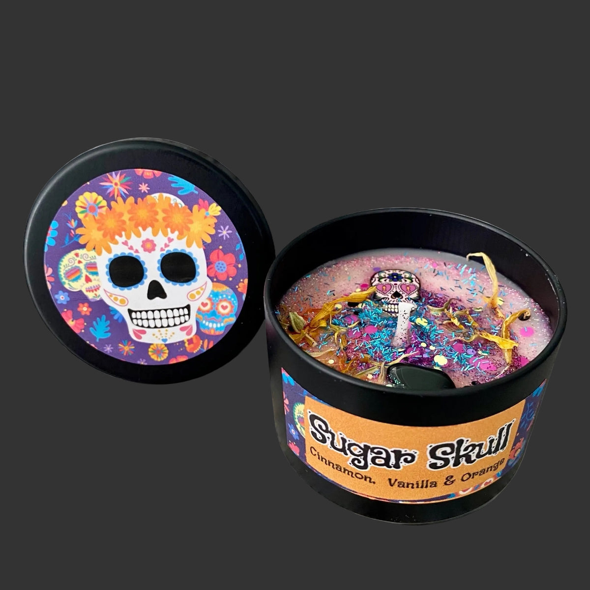 A candle in a black container with its lid placed beside it on a white surface. The lid features a colorful sugar skull design with flowers.