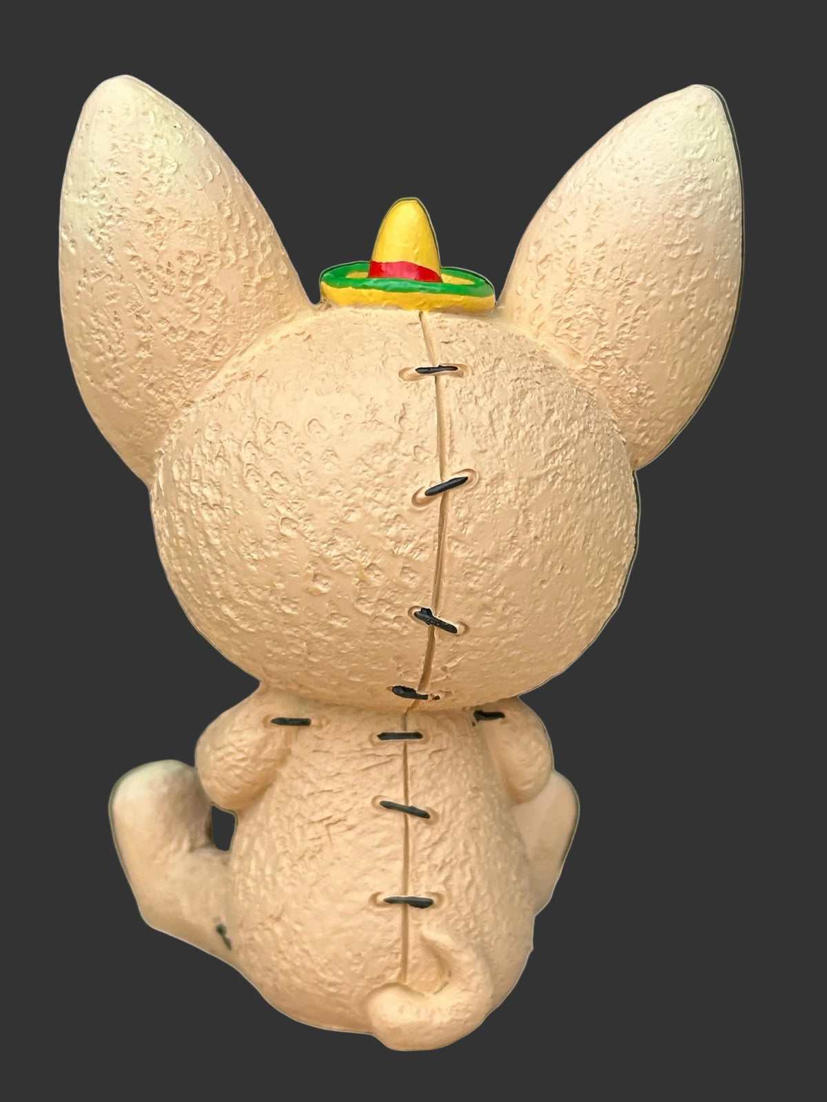 A beige textured  furrybones Chihuahua named Taco with large upright ears, black stitched seams, and a small colorful sombrero hat on top, shown against a plain black background.
