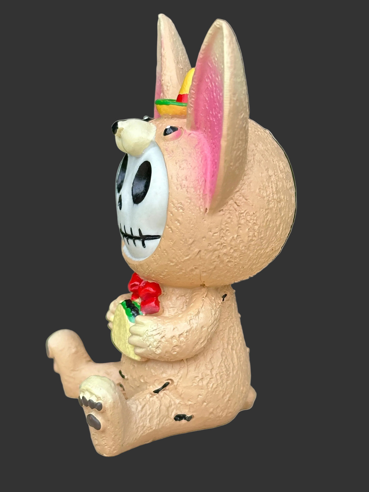 A beige textured Chihuahua  furrybones figurine with a skull face, holding a taco and wearing a red bow tie with a sombrero on his head on a plain black background.