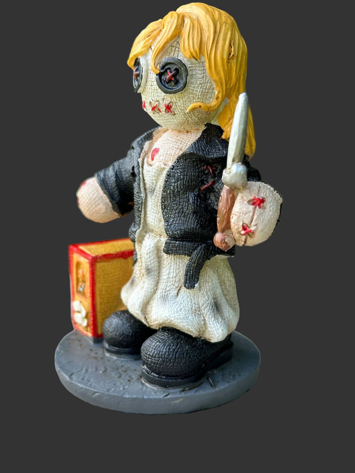 A painted horror-themed doll figurine resembling Leatherface, standing on a round black base. The figure has blonde hair, a stitched face with red stitched details, a black jacket, a cream-colored dress, and holds a knife in one hand while standing next to a small red and gold box.