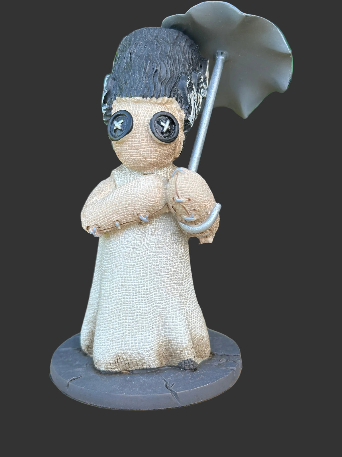 A handmade voodoo doll figurine with a beige linen-textured body, dark gray hair, button eyes, and a gray umbrella, displayed on a wooden surface with a blurred green garden background.