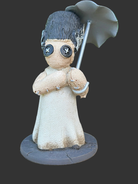 A handmade voodoo doll figurine with a beige linen-textured body, dark gray hair, button eyes, and a gray umbrella, displayed on a wooden surface with a blurred green garden background.