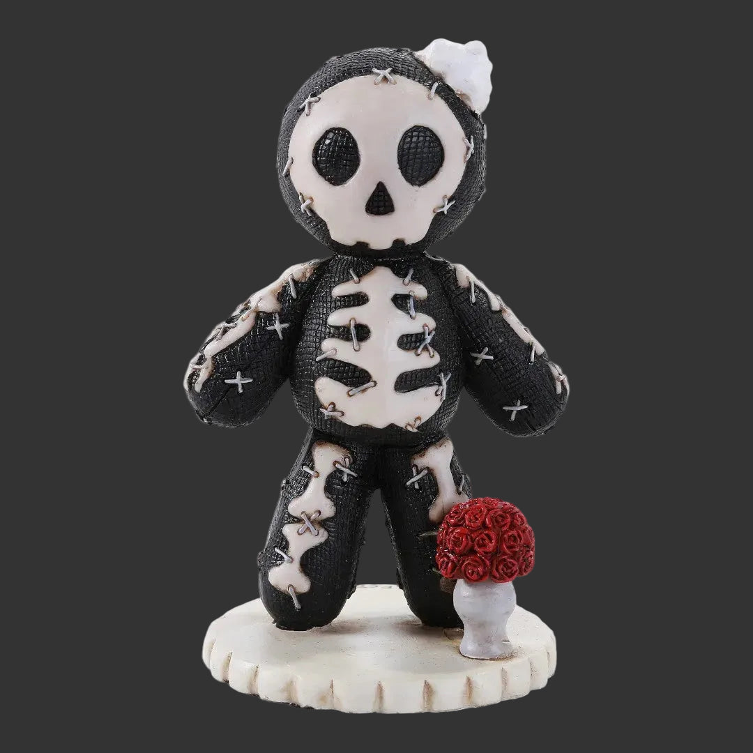 A gothic-style Pinhead Monster voodoo skeleton figurine with black and white stitched fabric texture, a small white flower on its head, and a red rose bouquet on a white pedestal base, isolated on a black background.