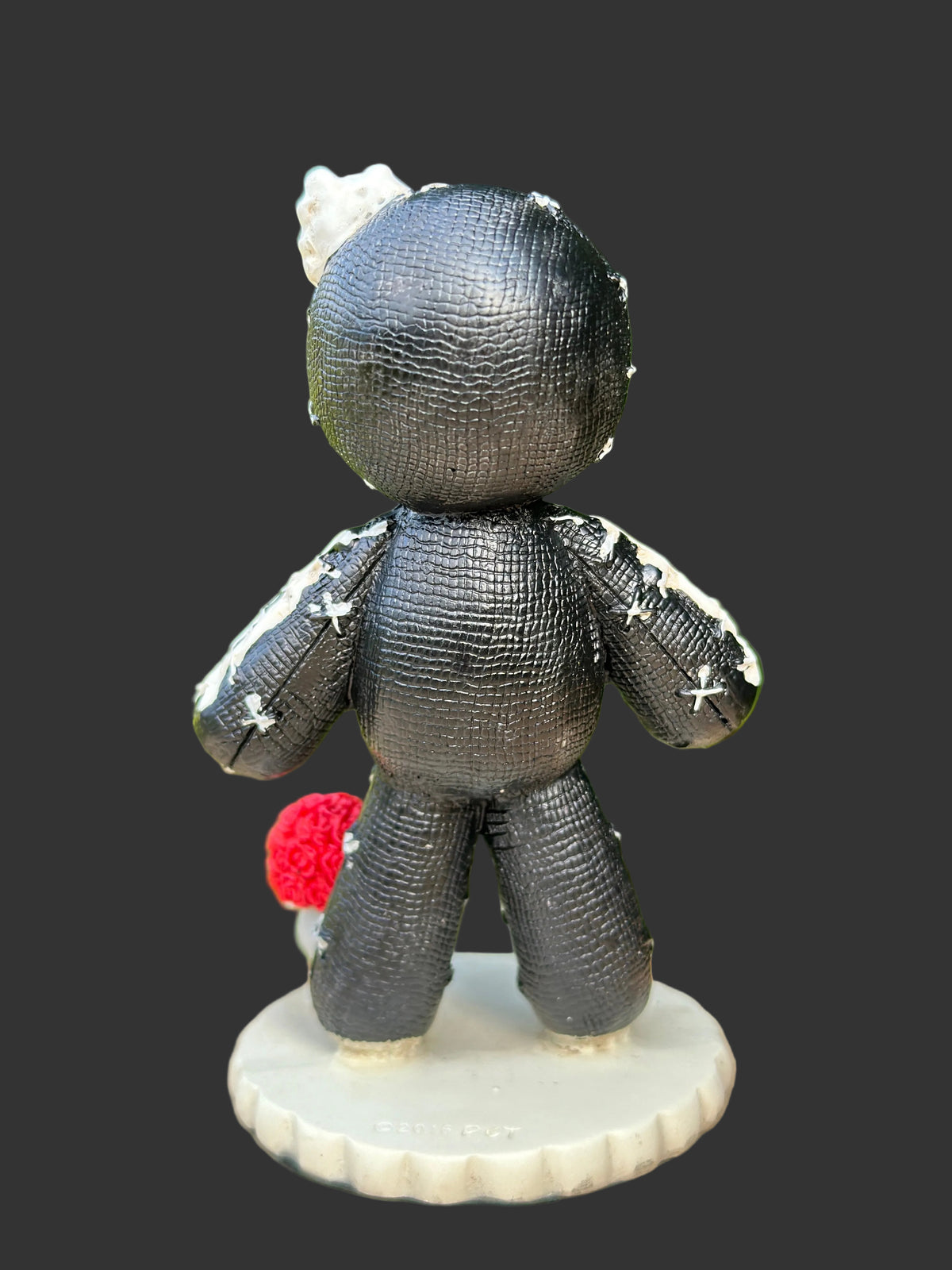 A small black stitched voodoo doll figurine shown from the back, standing on a round white base. The doll has white stitched details, a white flower in one hand, and a small red rose in the other.