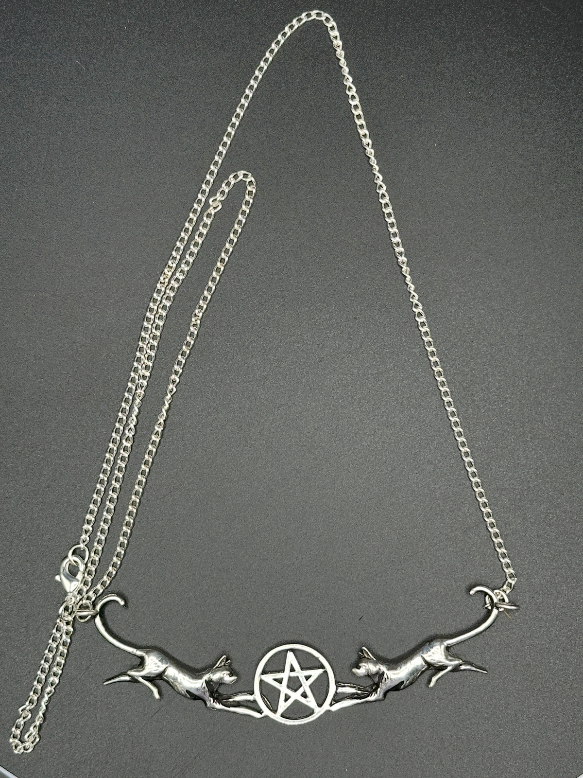 A silver-toned necklace featuring a central pentagram symbol flanked by two leaping cats, suspended from a curb chain on a dark gray background.