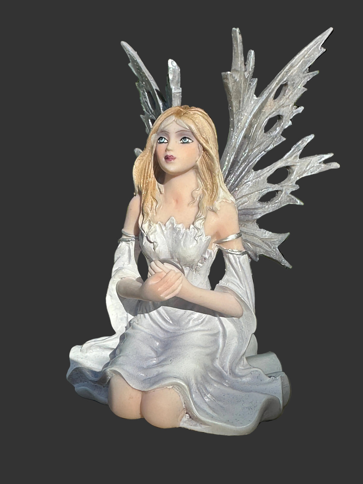 A kneeling Dream Eden winter fairy figurine with blonde hair, large translucent silver wings, and a white dress, shown against a dark gray background.