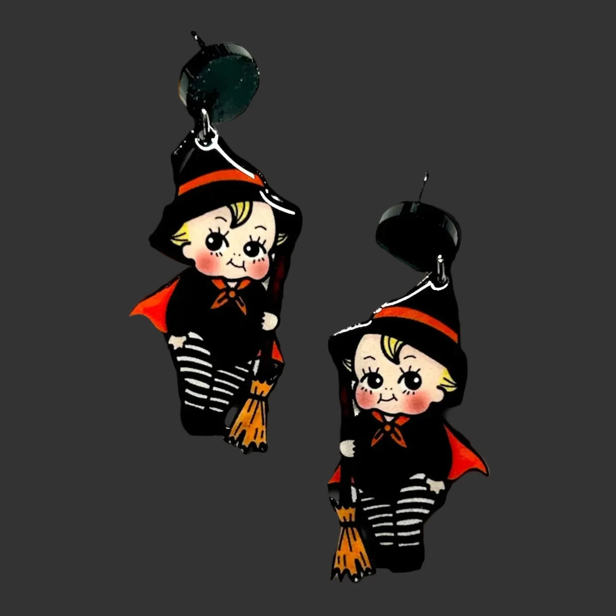 Two matching Halloween-themed dangling earrings featuring cartoon children in witch costumes, including black hats with orange trim, striped black-and-white pants, red capes, and orange broomsticks, shown against a black patterned background with crescent moons and stars.