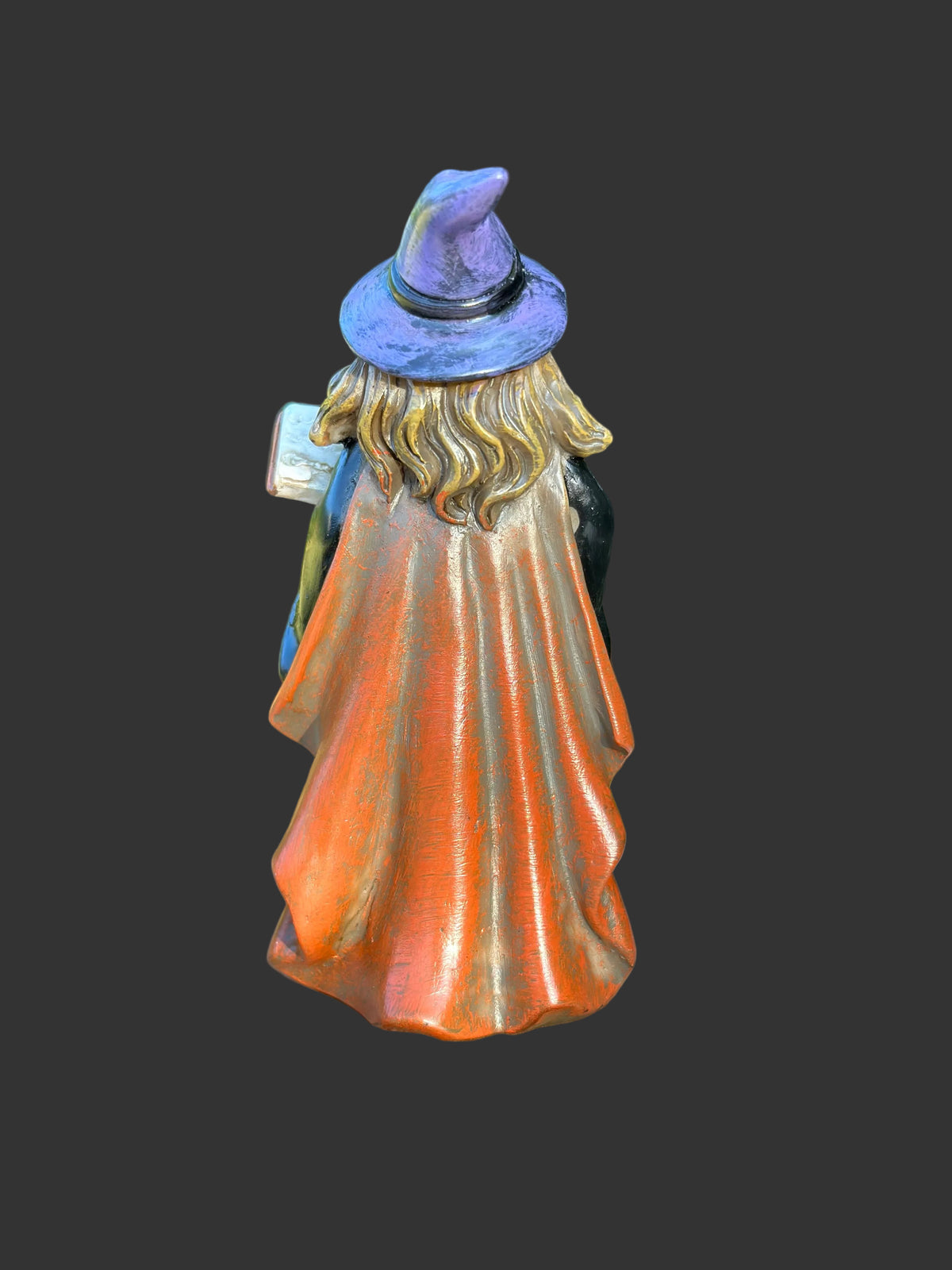 A miniature decorative figurine of a witch seen from the back, wearing a purple hat and a flowing orange robe with gold-toned highlights, standing on a cut tree stump with a blurred green garden background.