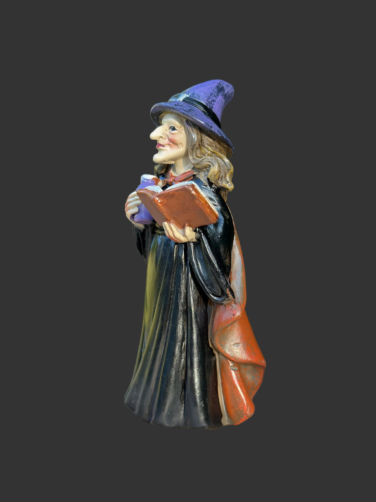 A resin miniature witch figurine with a purple hat, black robe, and orange-brown cloak, holding a book, displayed on a wooden surface with a blurred green garden background.