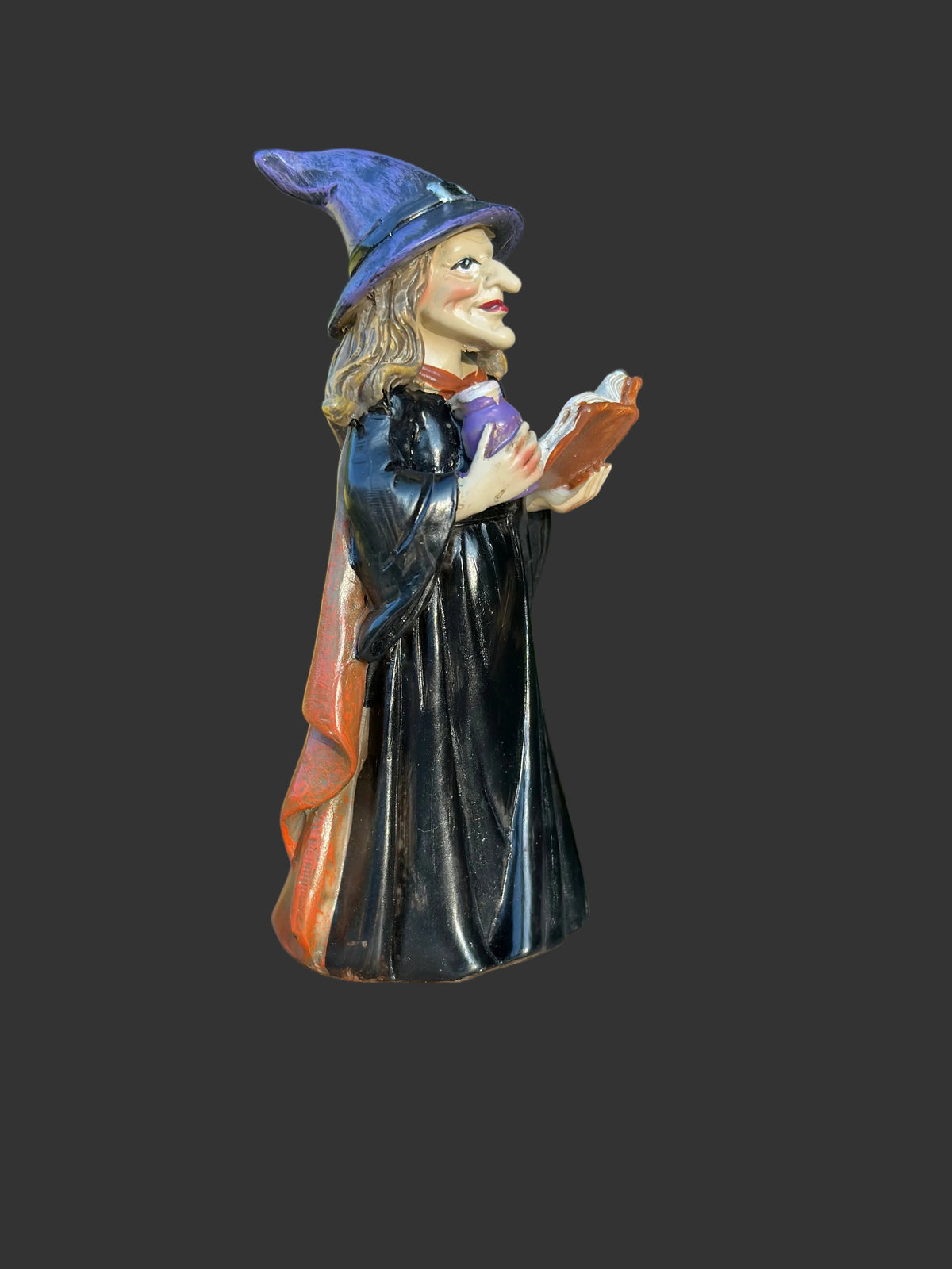 A small painted miniature witch standing on a round wooden surface with a blurred green garden background. The figurine wears a purple hat and black robe with an orange-red cape and holds an orange spell book.