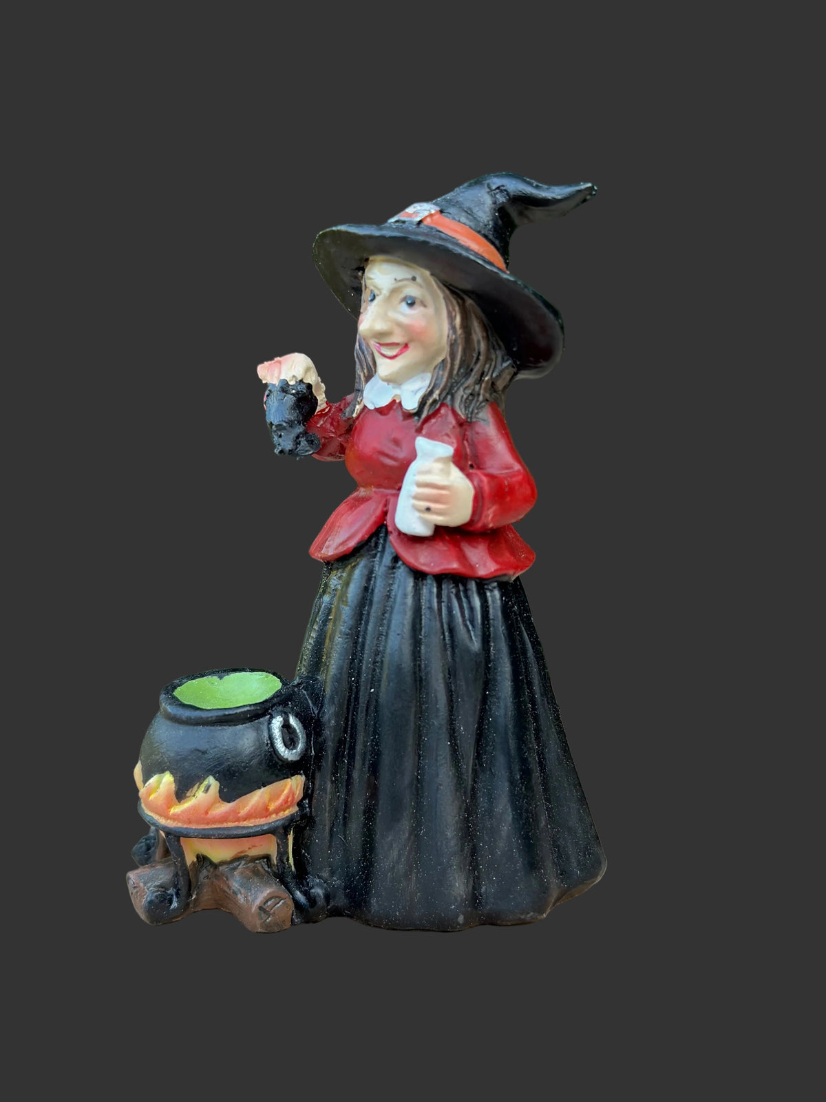 A small Halloween figurine of a witch in a black hat and skirt with a red top, standing beside a black cauldron with orange flames and a green liquid, placed on a wooden surface with a blurred green garden background.