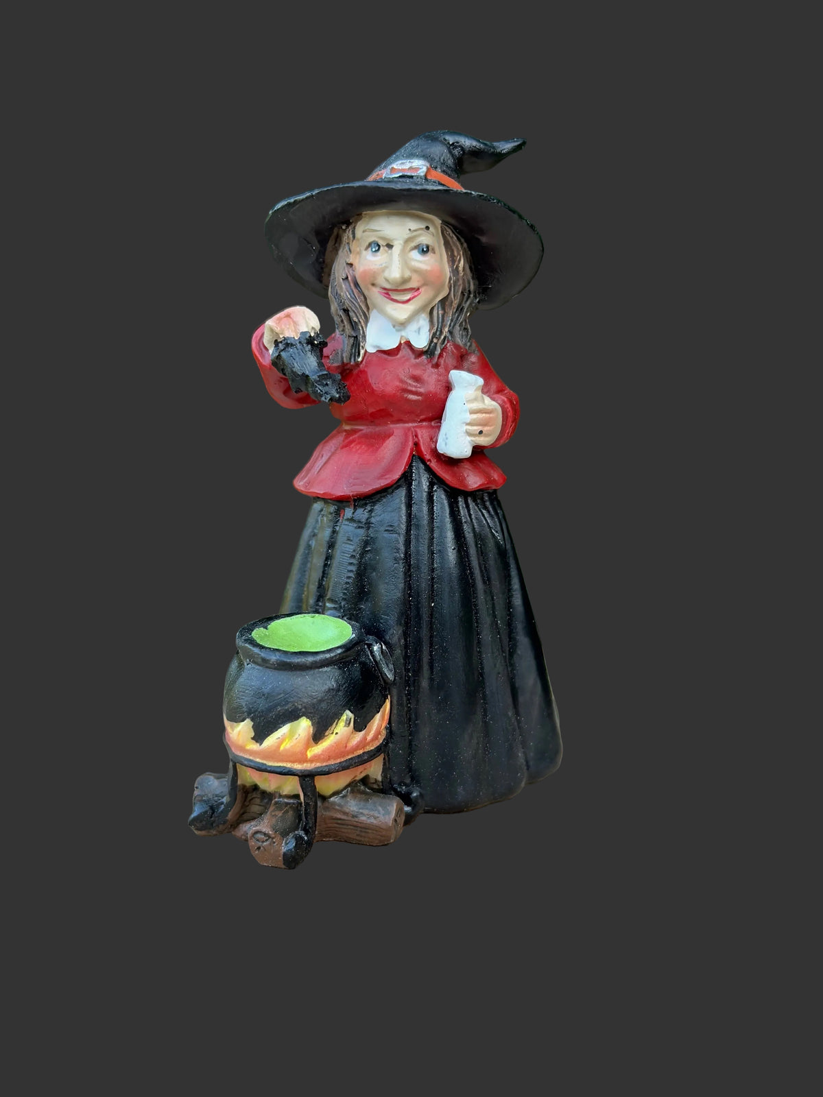 A miniature decorative witch figurine wearing a black hat and dress, holding a cauldron and potion bottle, displayed on a round wooden surface with a blurred green garden background.