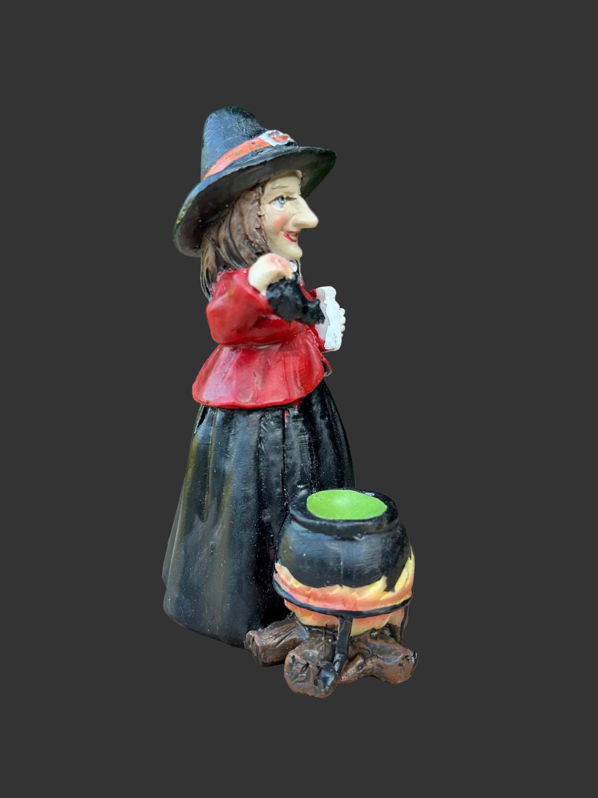 A small painted witch figurine stands on a circular wood slice with a blurred green garden background. The figurine wears a black hat and dress, a red top, and stands beside a black cauldron with a green contents and orange flames.