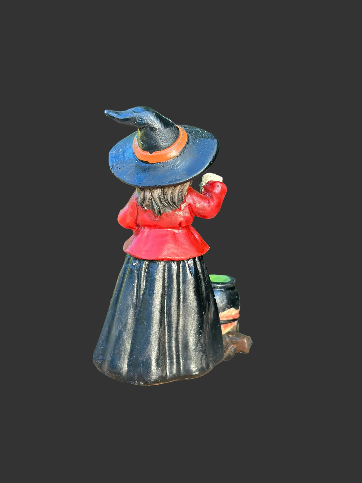 A small painted witch figurine shown from the back, wearing a black hat with an orange band, a red top, and a long black skirt, standing on a cut wood stump with a blurred green garden background.