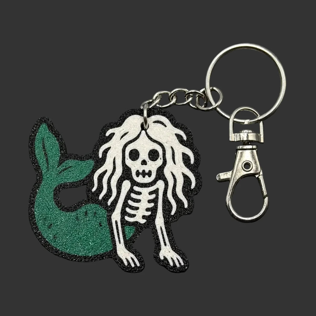 A mermaid and skeleton keychain charm with a green glitter mermaid tail and a white skeleton figure with long hair, attached to a silver metal key ring and clasp, displayed on a dark slate background.