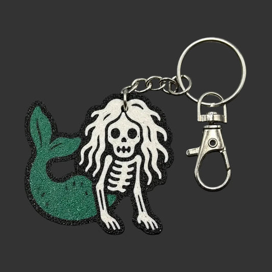 A mermaid and skeleton keychain charm with a green glitter mermaid tail and a white skeleton figure with long hair, attached to a silver metal key ring and clasp, displayed on a dark slate background.